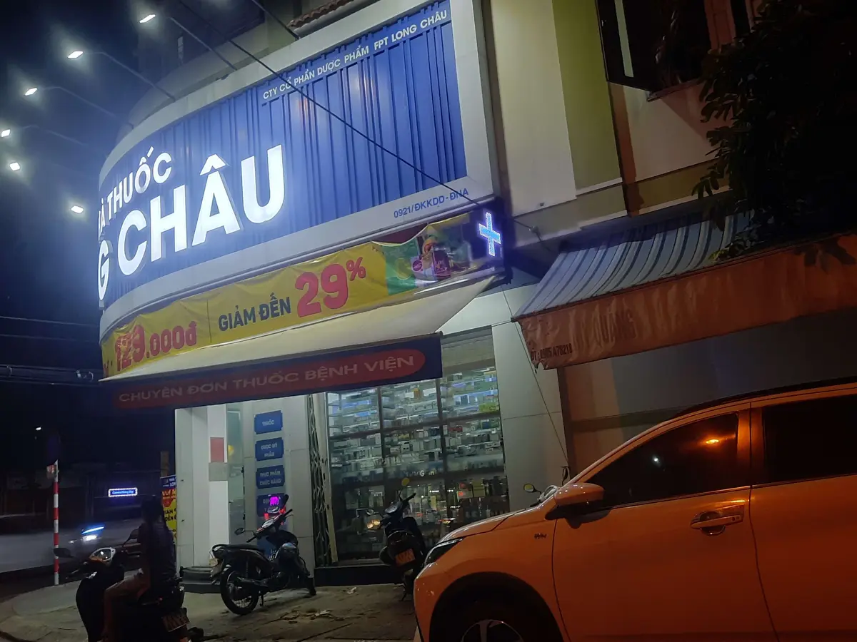 夜のLong Chau薬局の店先。青く光る看板、割引のバナー、停まっている2台のバイクが見える。