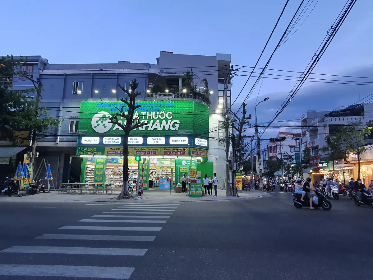 夕暮れのAn Khang薬局の角の店舗。通りの向かいから見ると、店先の上に緑色に光る看板がある。