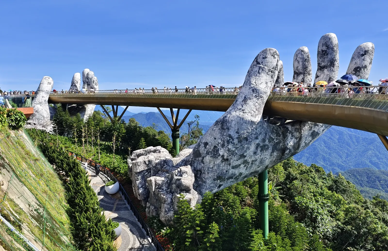 The Golden Bridge at Ba Na Hills, held up by two giant stone-look hands, crowded with visitors under umbrellas.