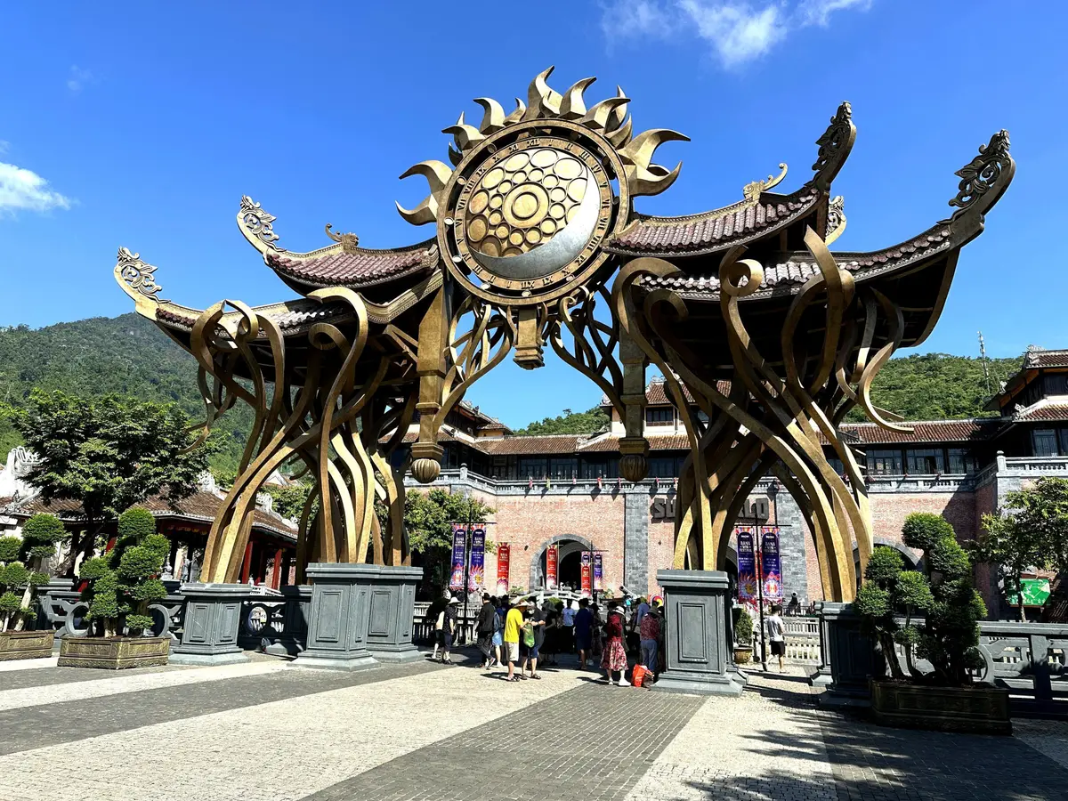 一座雕刻精美的入口大门,图案似日月,后面是红瓦的寺庙式屋顶线条,游客从门下走过。