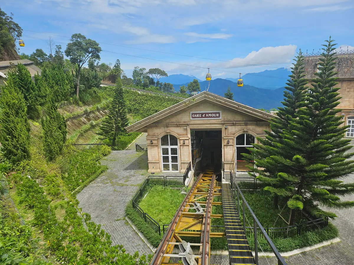 一座小型铁轨车站建筑,轨道通向车站,缆车吊厢从头顶经过,背景是群山。
