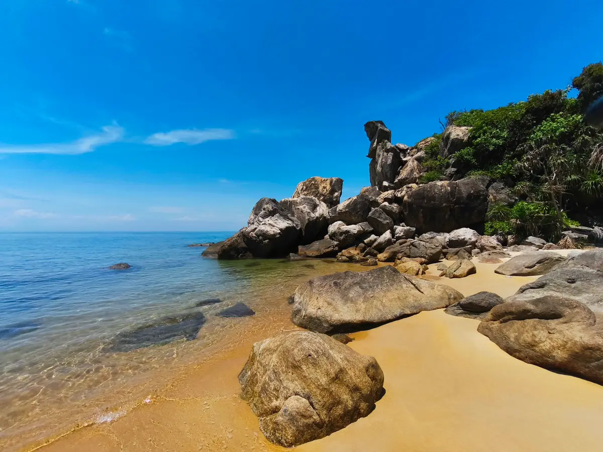 A golden beach with large boulders at the water's edge and clear, shallow sea under a blue sky.
