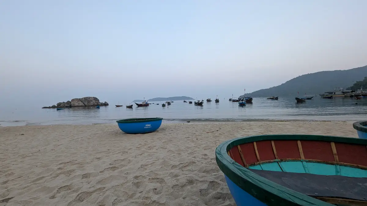 Round coracle boats on the sand and fishing boats at anchor on a calm sea in early light.