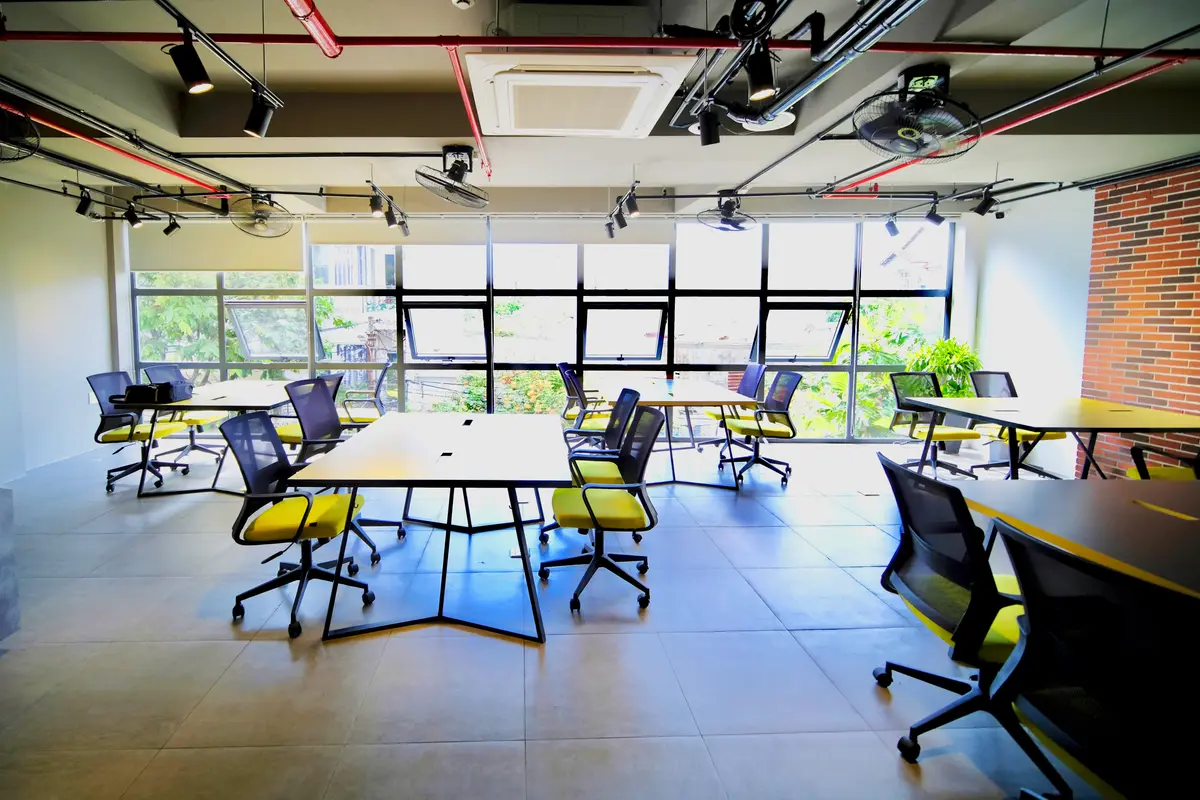 Ein Raum mit langen Tischen und Netzstühlen mit gelben Sitzflächen vor einer großen Fensterfront, mit freiliegenden Deckenrohren, Schienenstrahlern und Backsteinwand.