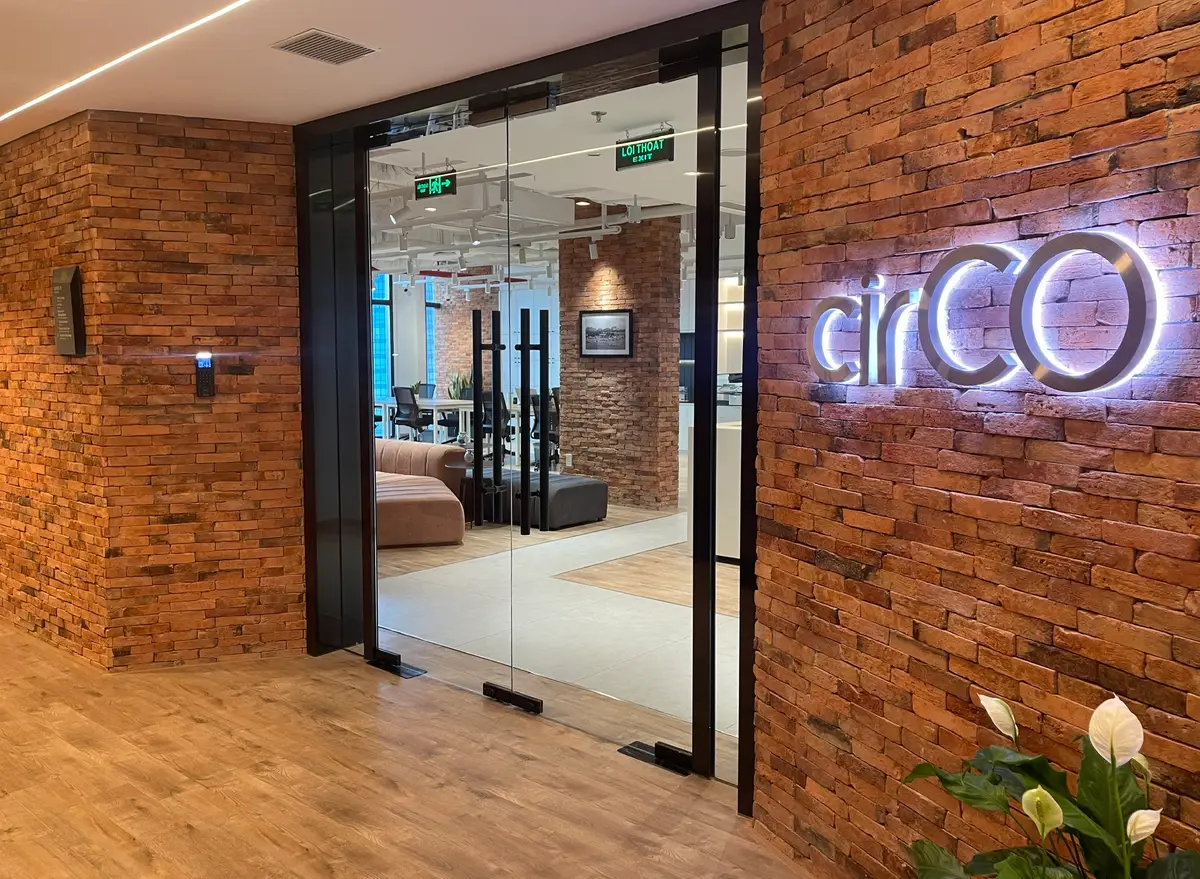 Eine Glastür in einer roten Backsteinwand, an der Wand ein beleuchteter cirCO-Schriftzug, hinter der Tür eine helle Coworking-Fläche mit Sofa und Schreibtischen.
