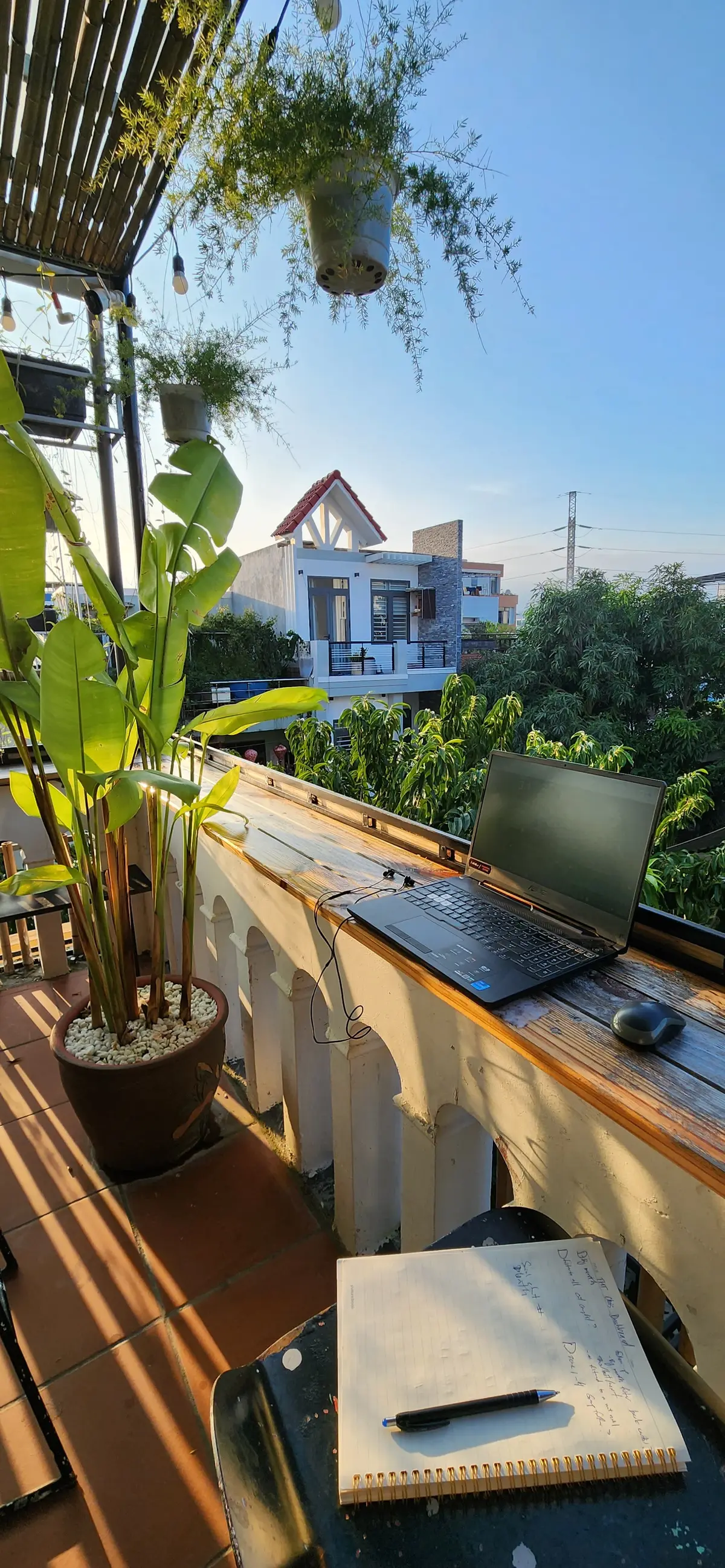 Ein Laptop und eine Maus auf einer hölzernen Stehtheke am Balkongeländer, mit Hängepflanzen, einer Topfpalme und Nachbarhäusern im Hintergrund.