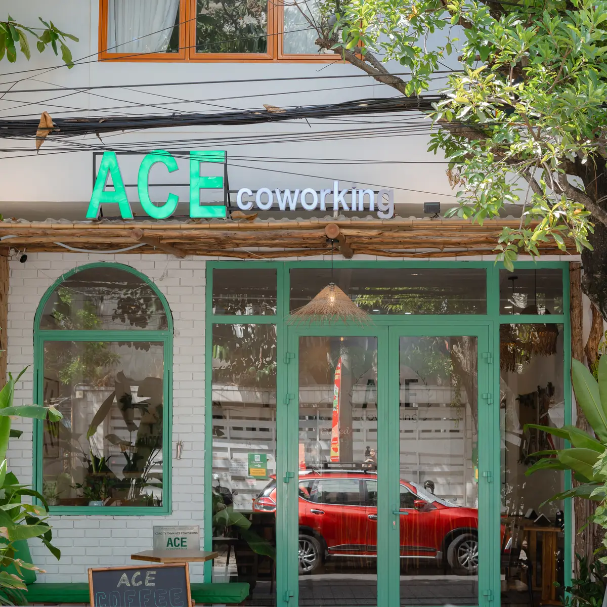 Die Ladenfront von ACE Coworking mit grün leuchtendem ACE-Schriftzug, grün gerahmten Rundbogenfenstern, weißer Backsteinwand und einer Kreidetafel an der Tür.