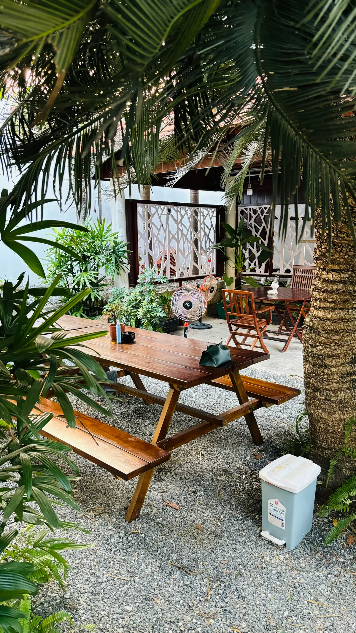 Unter Palmwedeln in einem Kiesgarten stehen ein Picknicktisch aus Holz und Klappstühle, dahinter zwei Standventilatoren und ein gemusterter Sichtschutz.