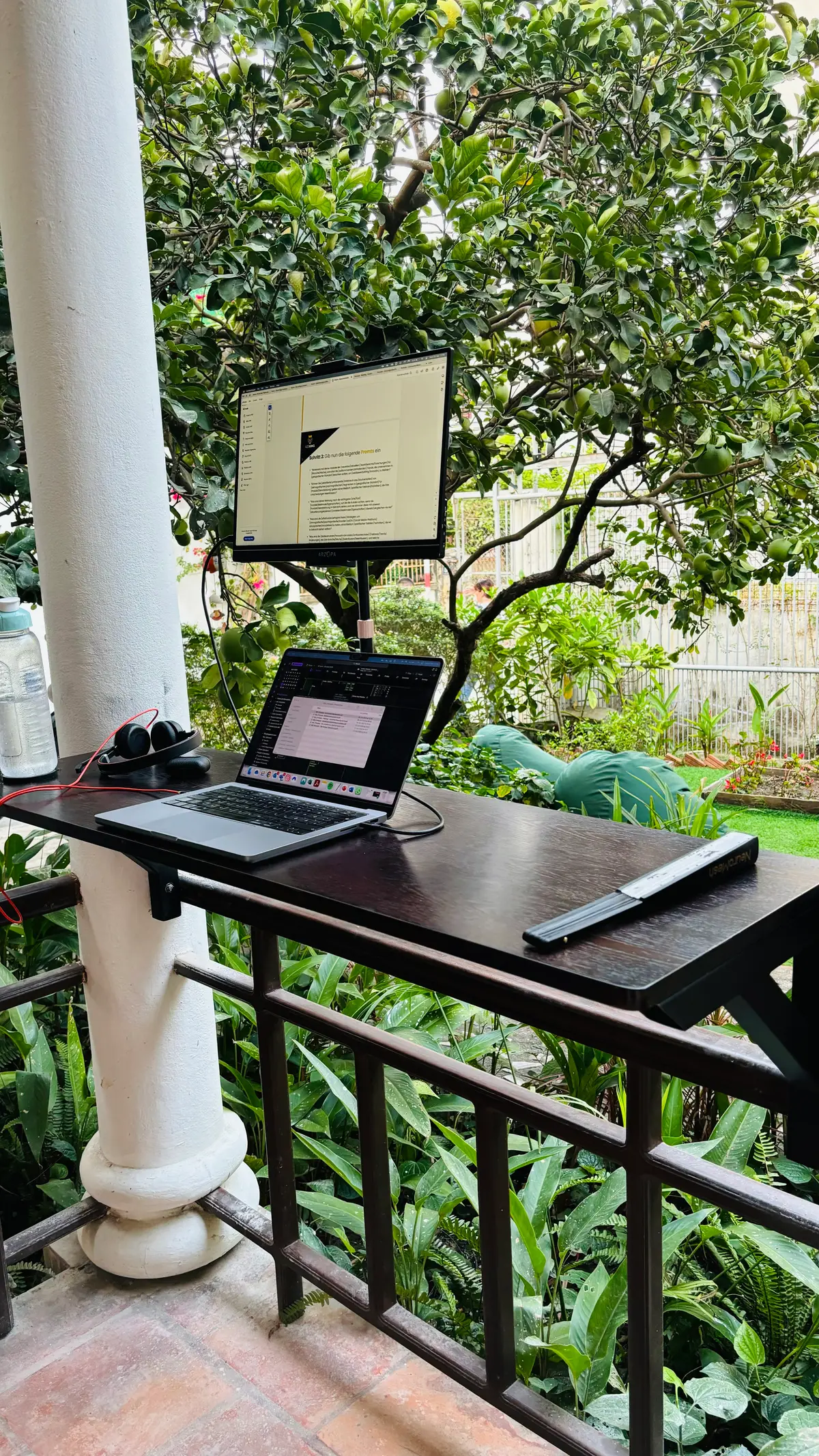 Ein dunkler Holzschreibtisch auf einem überdachten Balkon mit Laptop, erhöhtem Monitor und Kopfhörern, umgeben von Gartenpflanzen und grünen Sitzsäcken.