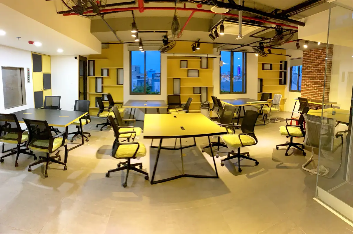Ein leerer Coworking-Raum mit schwarzen und gelben Tischen, Netzstühlen mit gelben Sitzflächen, gelben Regalen, Backsteinwand und Glastrennwand.