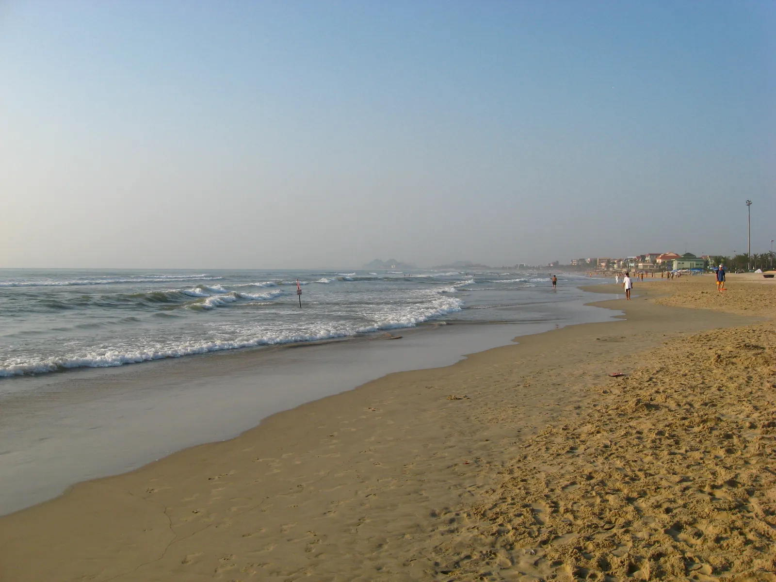 Mỹ Khê beach in Da Nang on a clear morning: waves breaking along a wide sandy shore, a few people walking at the water's edge and hotels along the seafront in the distance.