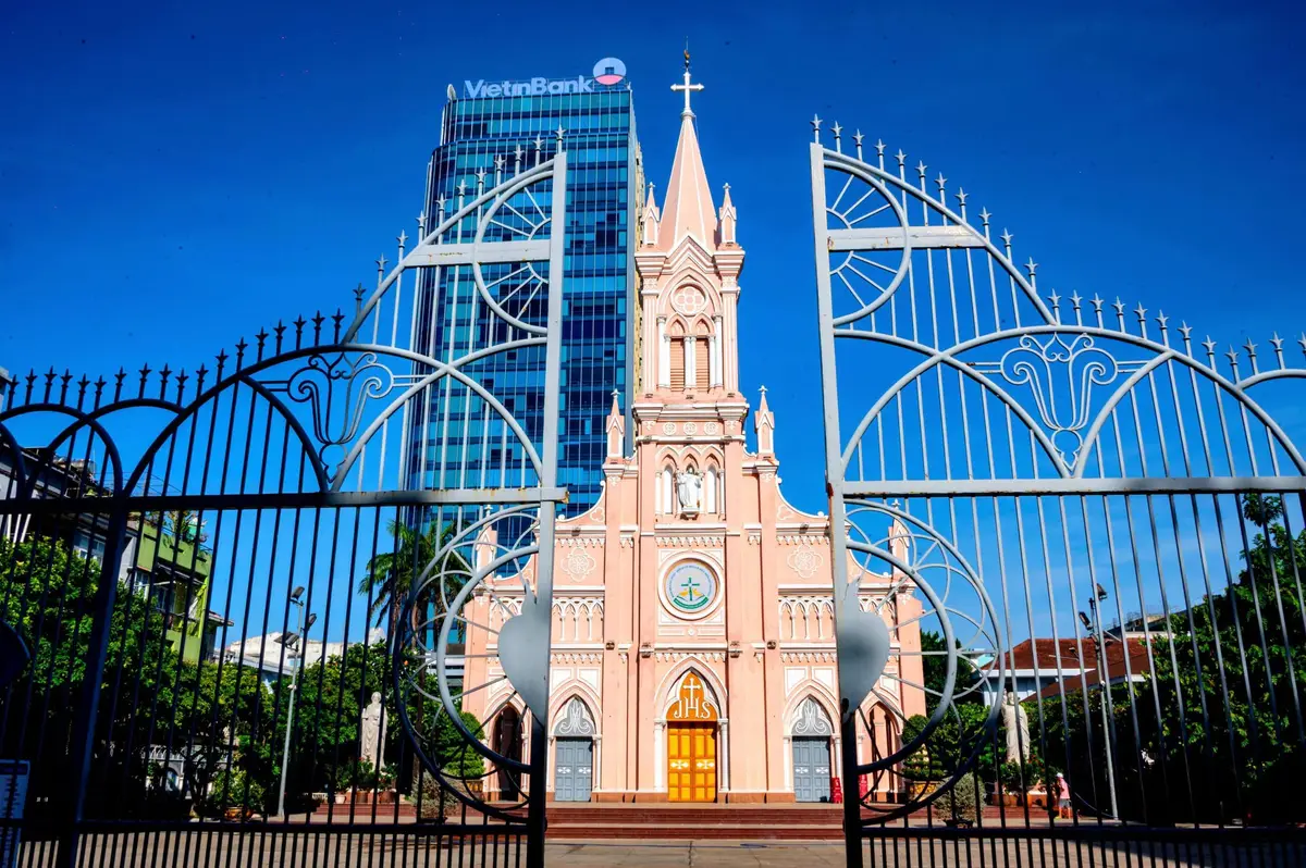 The pink Gothic front of Da Nang Cathedral seen through its open iron gates on Tran Phu Street, with a glass office tower behind.