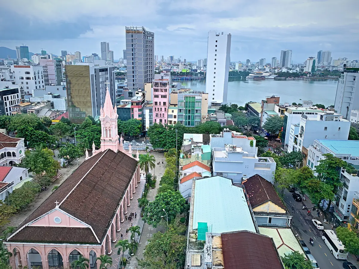 Aerial view of Da Nang Cathedral's long pink nave and spire among city buildings, with the Han River behind.