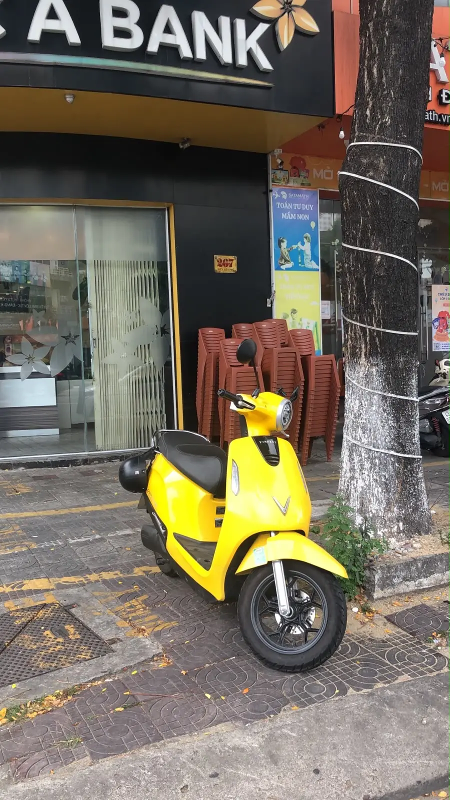 一辆黄色踏板摩托车停在路缘，旁边是一棵树和一家店面。