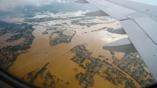 Aerial view from an aircraft window of wide brown floodwater covering fields and villages, with settlements standing out of the water