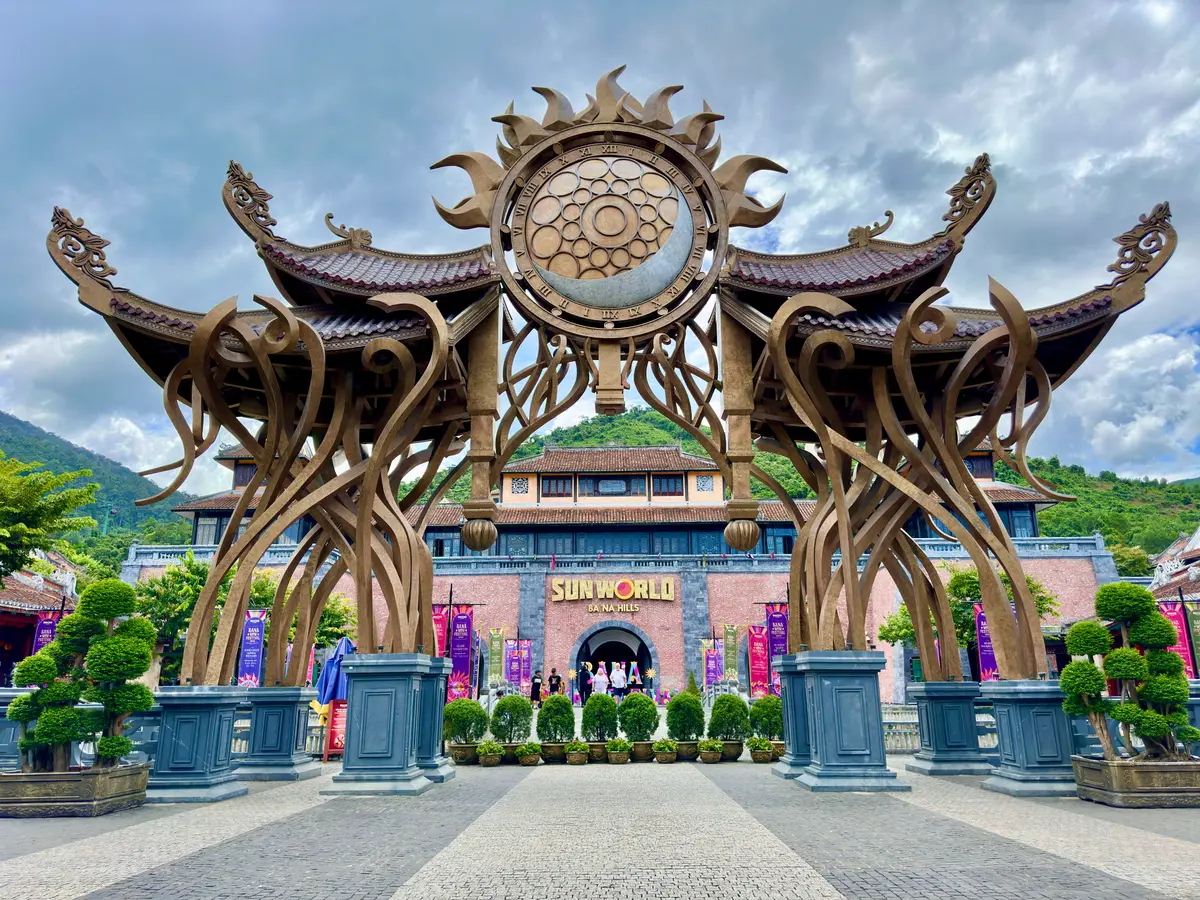 Sun World Ba Na Hills 입구 구역. 포장된 광장 위에 금색 태양 문양이 있는 화려한 문이 서 있어요.