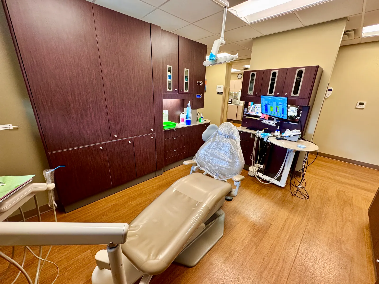An empty dental treatment room with a reclined dental chair under a plastic cover, a light arm, cabinets and a computer screen.