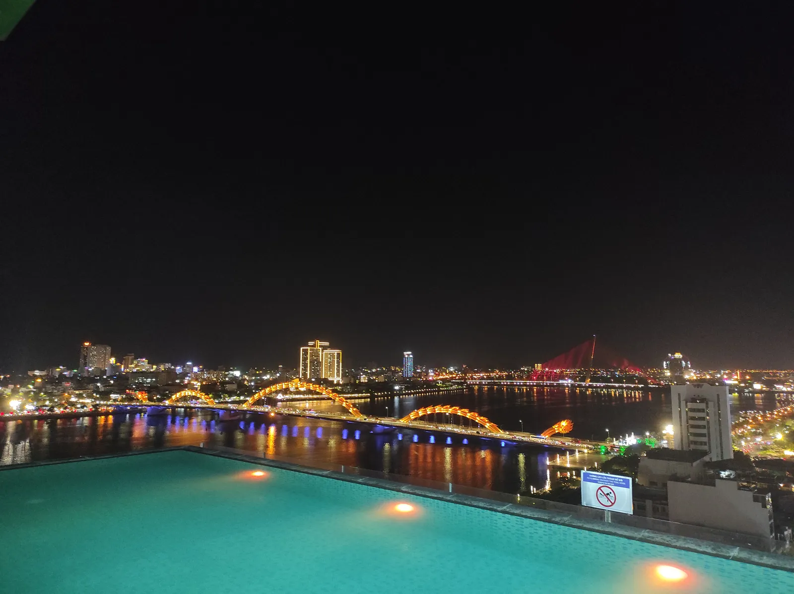 Night view over the Han River from a rooftop pool: the Dragon Bridge lit in gold along its arches, with the city lights and a red-lit cable bridge further upstream.