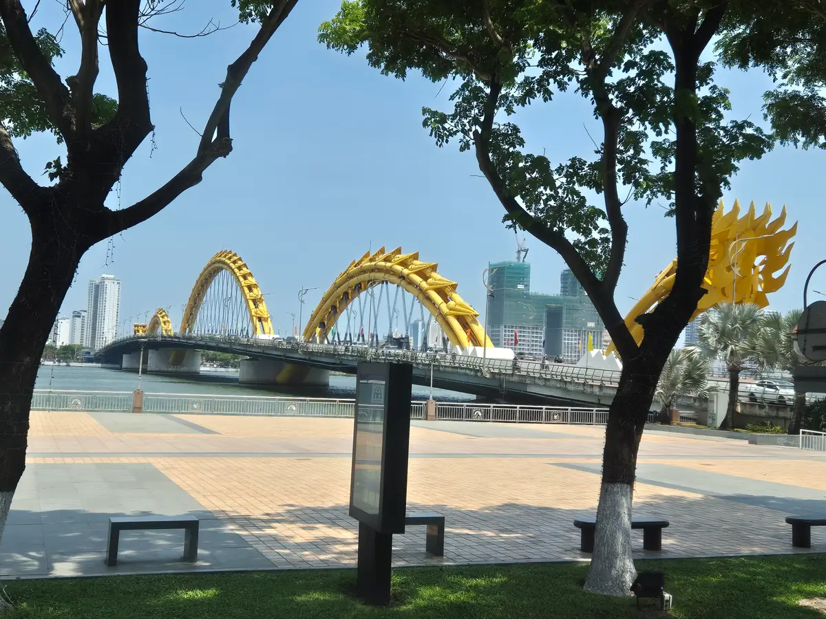Tagesansicht der goldenen Brückenbögen auf der anderen Flussseite, eingerahmt von Bäumen an einer gepflasterten Uferpromenade.