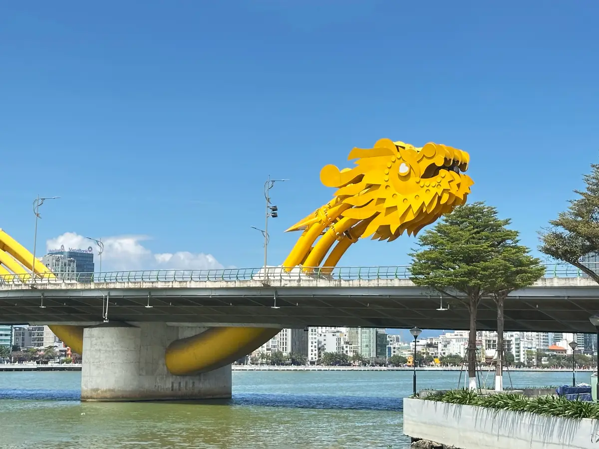 Nahaufnahme des goldenen Drachenkopfs bei Tag: Er erhebt sich vor wolkenlosem blauem Himmel von der Fahrbahn der Brücke.