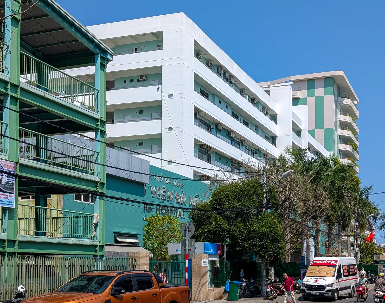 Da Nang Hospital seen from Hai Phong Street: white and green ward buildings behind the hospital's gate, with an ambulance and motorbikes on the street in front.