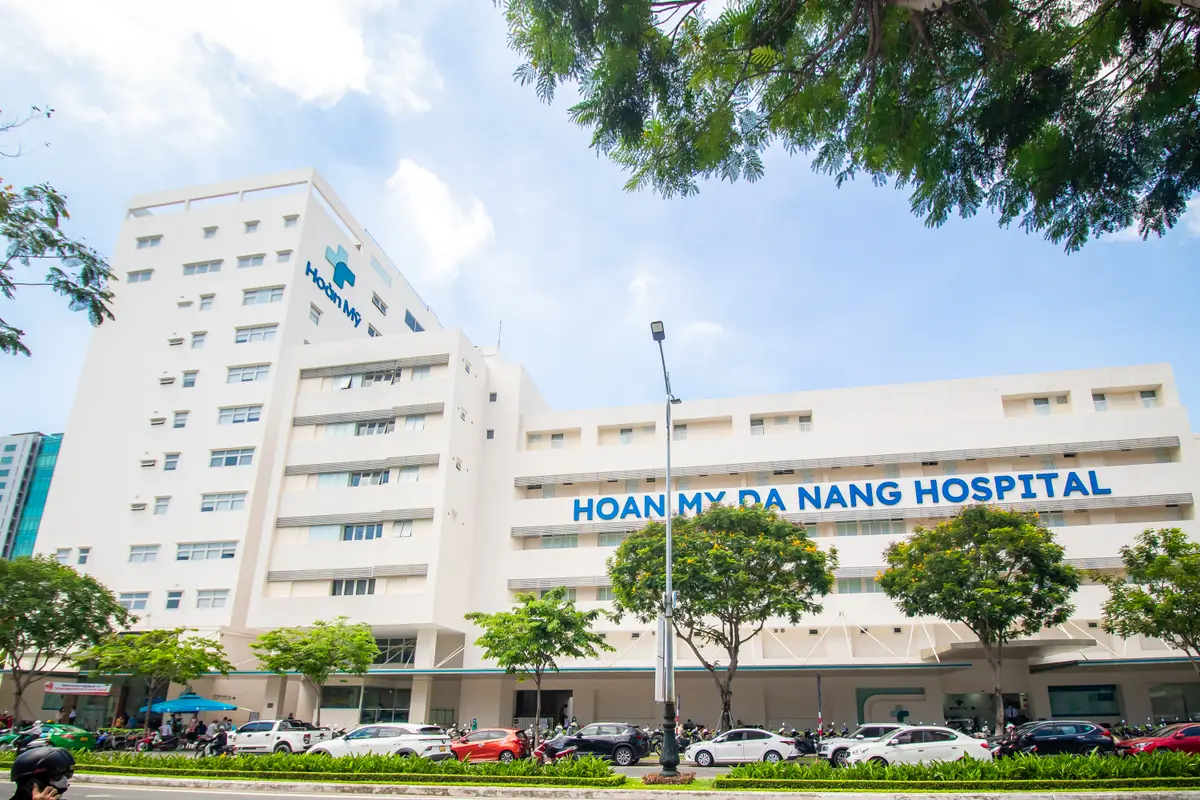 파란 글씨로 이름이 적힌 흰색 Hoan My Da Nang Hospital 건물. 앞에 나무와 주차된 차들이 있어요.