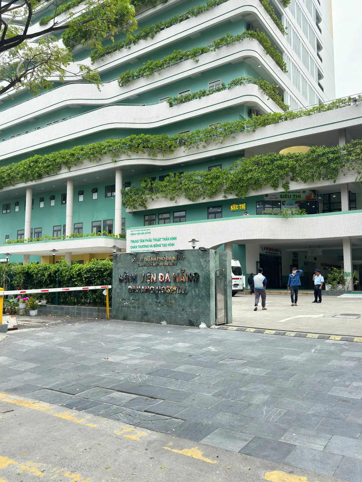 흰색과 초록색 건물 앞에 있는 Da Nang Hospital의 정문과 이름 간판. 건물을 덩굴 식물이 덮고 있어요.