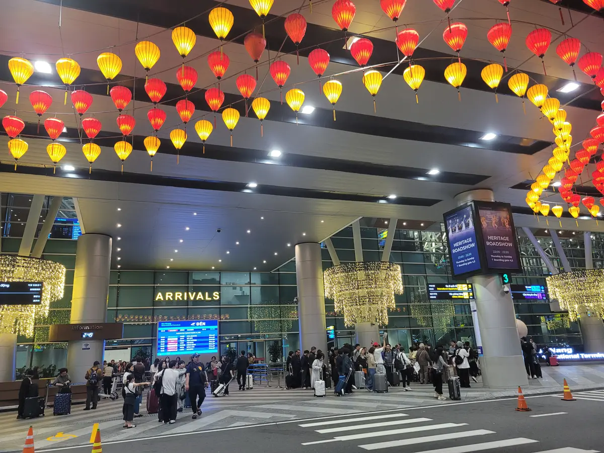 夜晚的到达厅正面，头顶挂着灯笼，有“ARRIVALS”标牌，人行道上有带着行李的旅客。