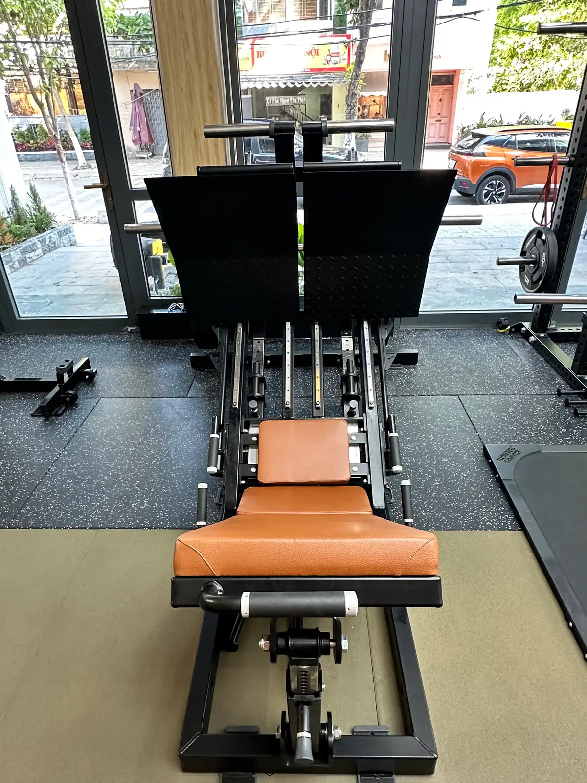 Leg press machine and racks on a rubber gym floor beside large windows