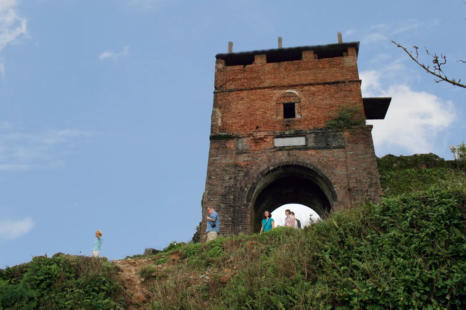 The brick gate tower of Hải Vân Quan on a grassy hilltop, with a few visitors walking through the arch.