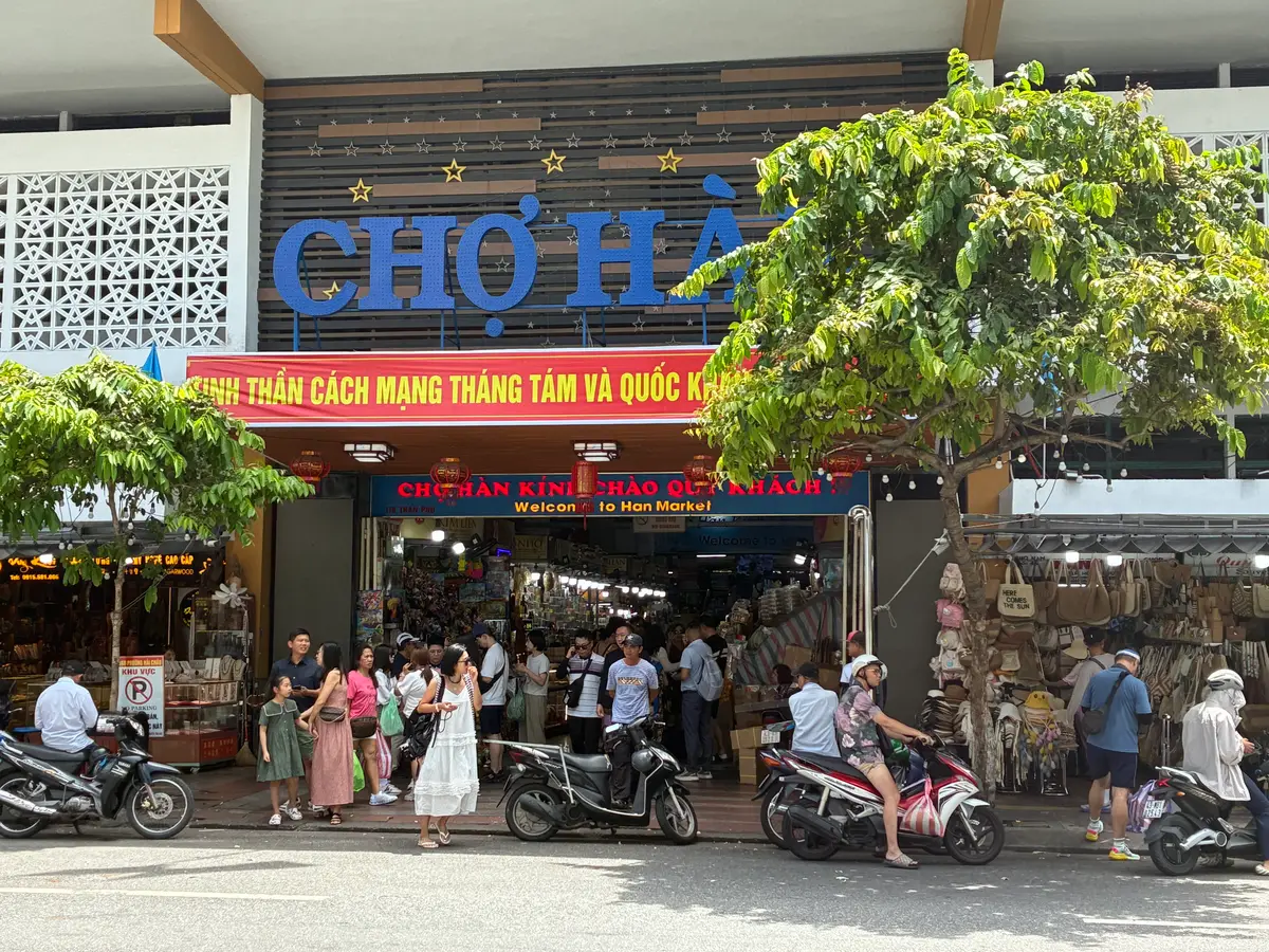 The front of Han Market with its large blue market sign, motorbikes parked on the pavement and shoppers at the entrance.