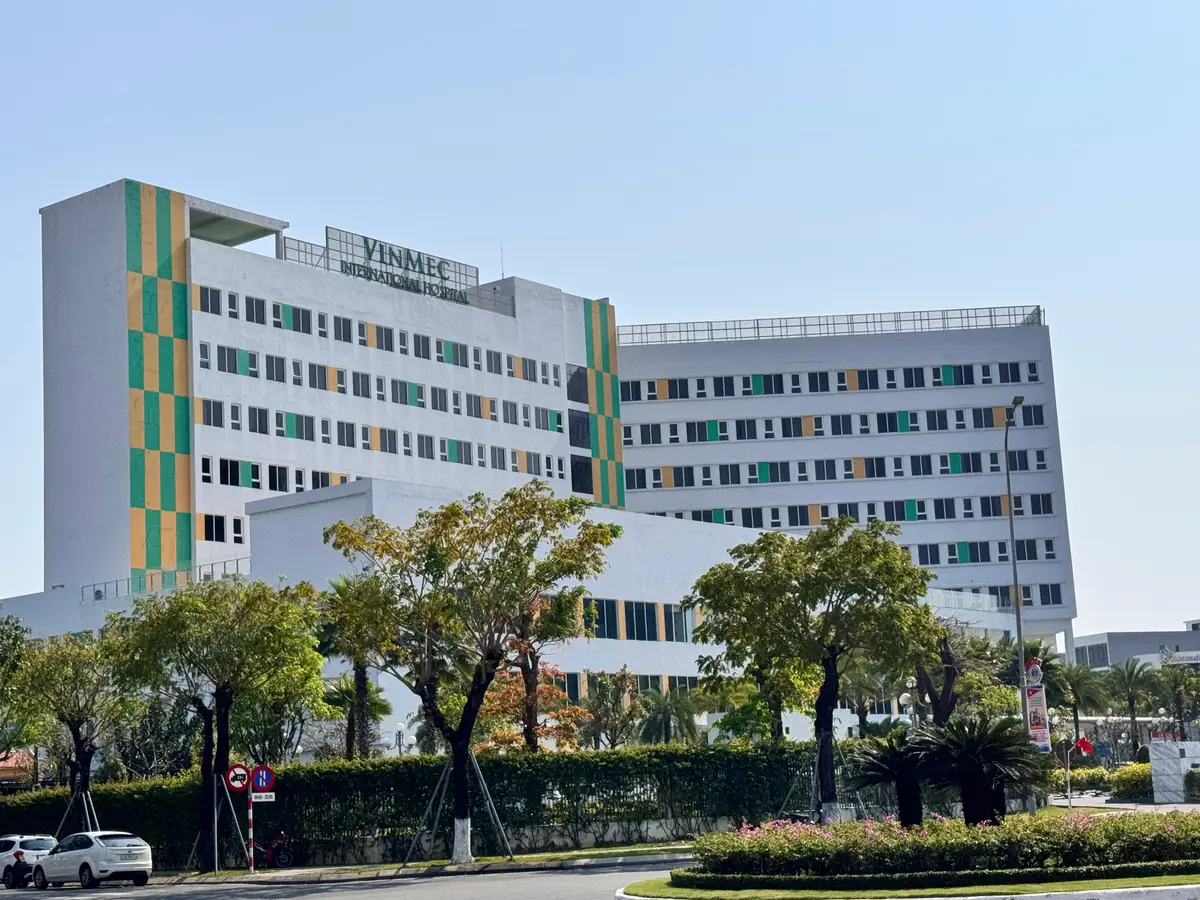 잔디밭과 나무 너머로 보이는, 초록색과 노란색 패널이 있는 흰색 Vinmec International Hospital 건물.