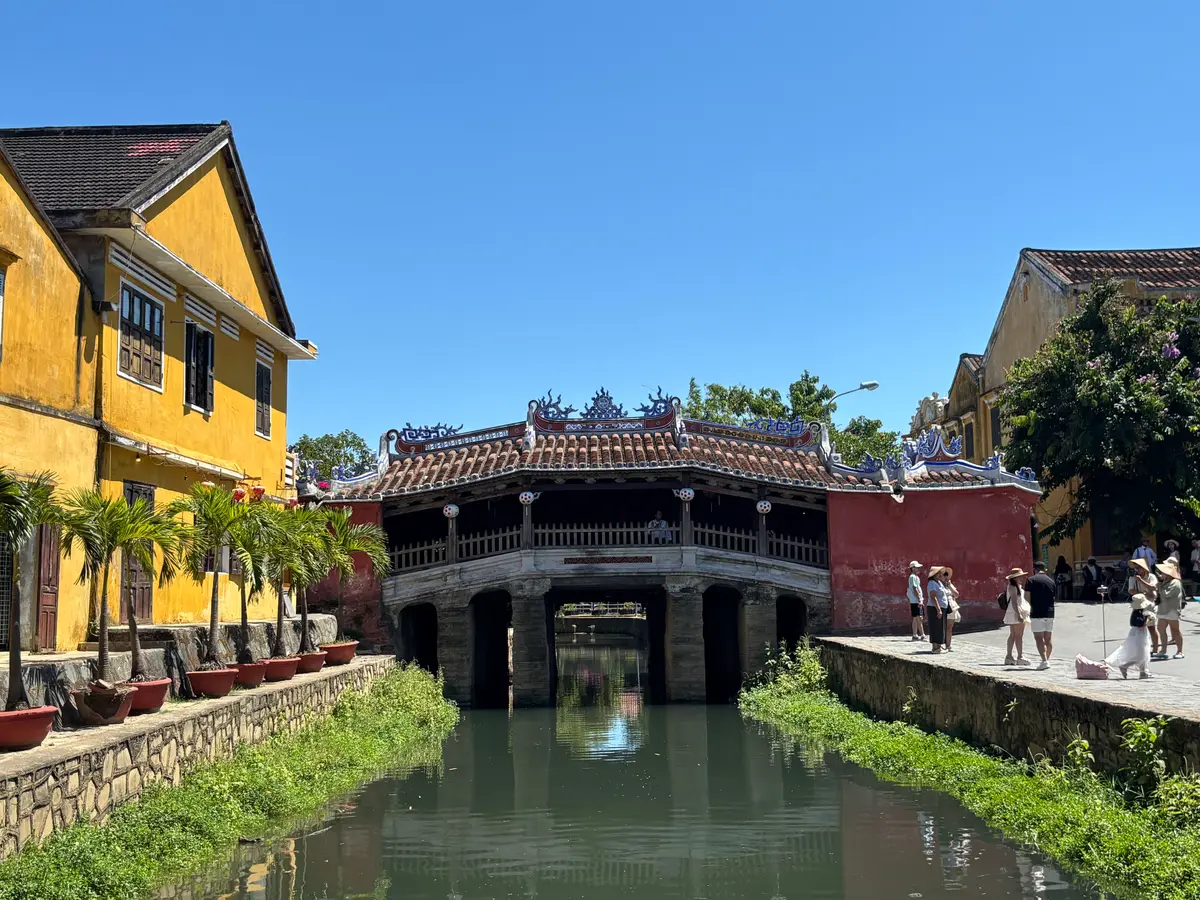 Die Japanische Brücke wölbt sich über einen Kanal, ihr Ziegeldach trägt geschnitzte Drachenmotive; daneben stehen gelbe Handelshäuser, am nahen Ufer Besucher.