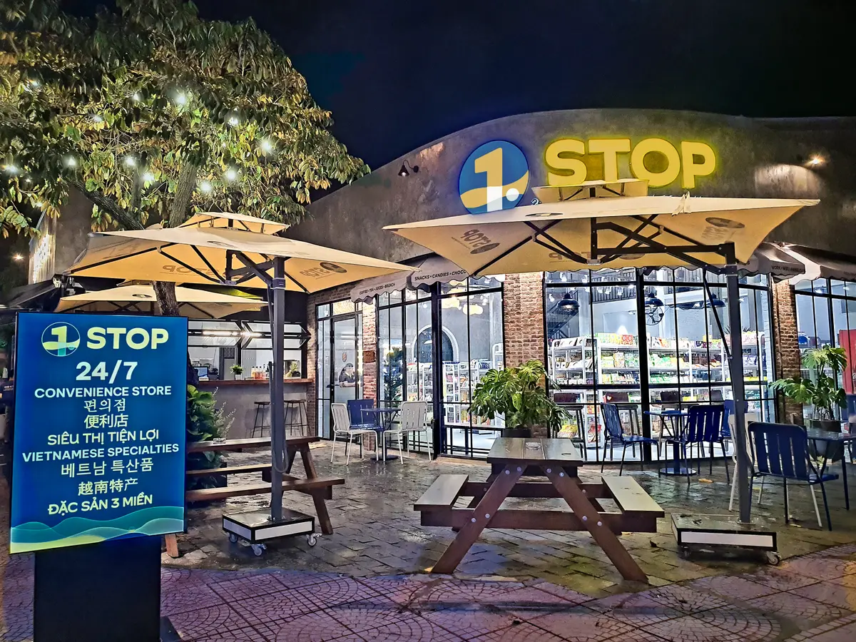 Glass-fronted convenience store at dusk with a lit 1 STOP sign and outdoor umbrellas