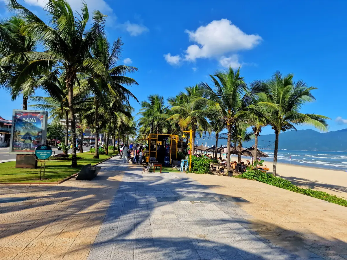 A paved promenade lined with palm trees beside My Khe Beach, with the sea and hills in the distance.