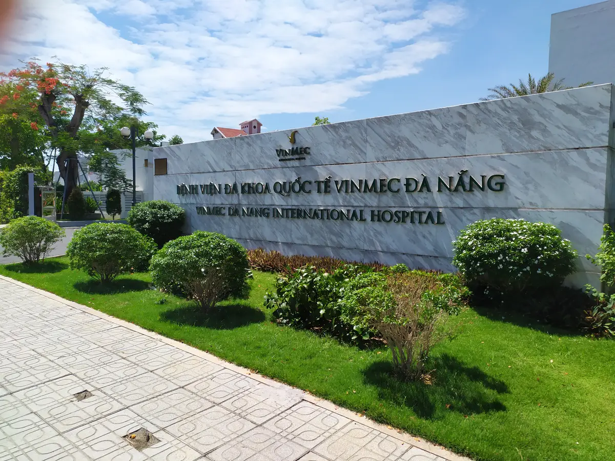 A grey marble entrance wall with the Vinmec Da Nang International Hospital name in dark letters, behind a lawn and shrubs.
