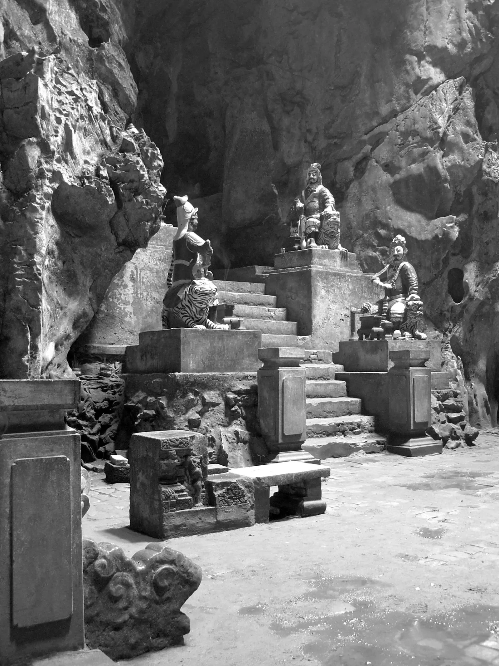 Black-and-white photo of a cave shrine in the Marble Mountains: painted guardian statues on stone plinths and steps against the rock wall.