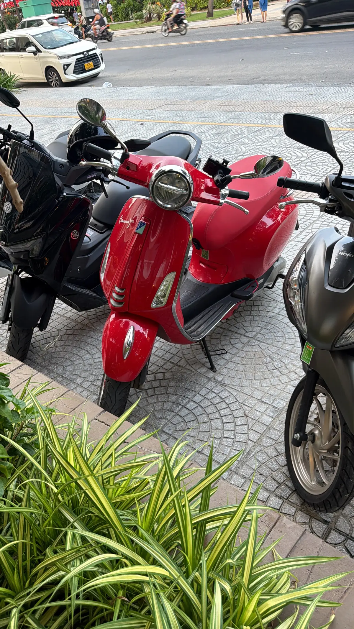 Ein roter Roller im Vespa-Stil steht auf einem gepflasterten Gehweg zwischen einem schwarzen Roller und einer grauen Honda.
