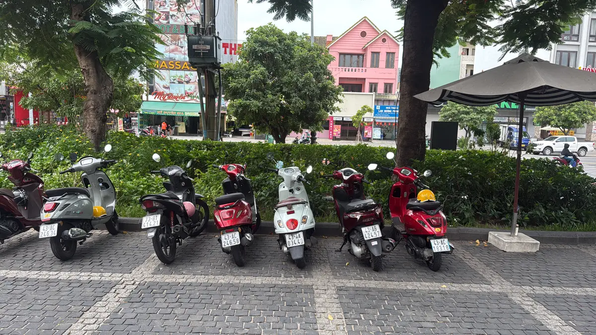Eine Reihe aus sechs Rollern in Rot, Schwarz, Grau und Weiß steht vor einer Hecke an einer gepflasterten Straße.