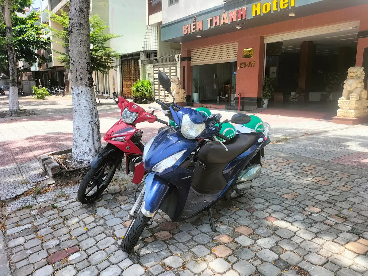 Ein blauer Honda-Roller und ein rotes Zweirad mit grünen Helmen auf dem Sitz stehen auf einem gepflasterten Gehweg vor einem Hotel.