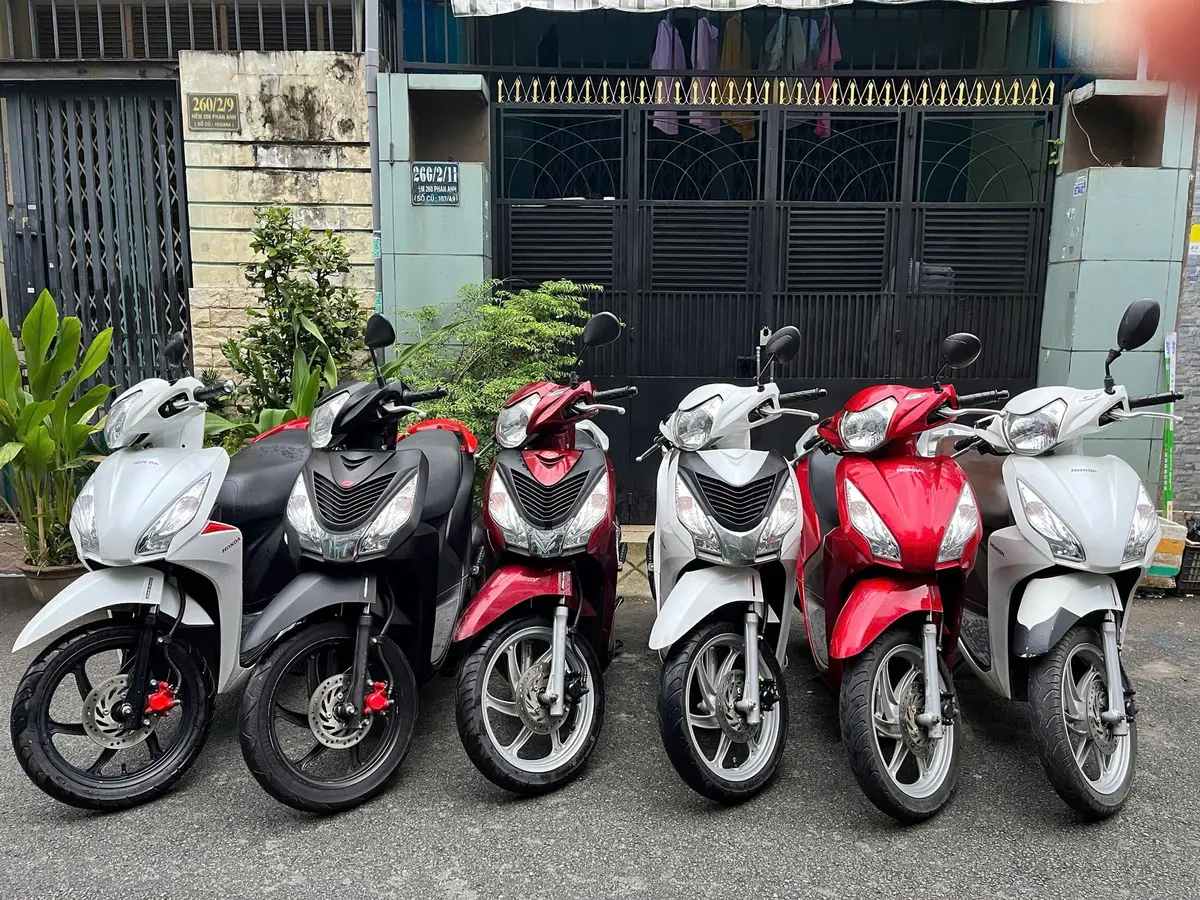 Sechs Honda-Roller in Weiß, Schwarz und Rot stehen in einer Reihe vor einem schwarzen Tor in einer schmalen Straße.