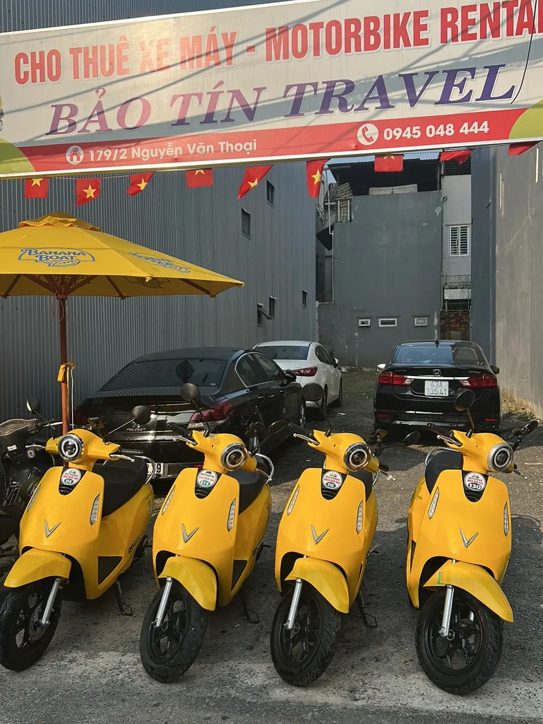 Vier gelbe Elektroroller stehen in einer Reihe unter einem großen Schild mit der Aufschrift „Bao Tin Travel motorbike rental“.