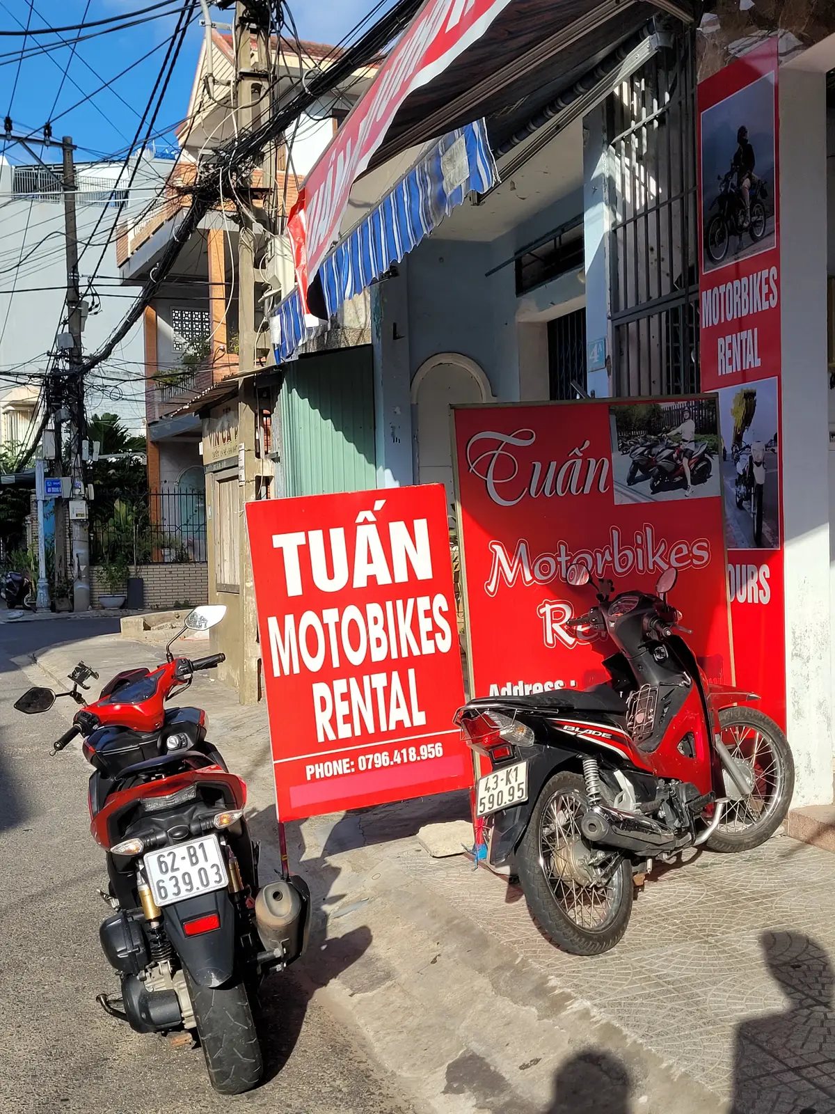 Rote Schilder von Tuan Motorbikes Rental an einer Straße in Da Nang, davor stehen ein roter Roller und eine rote Honda.