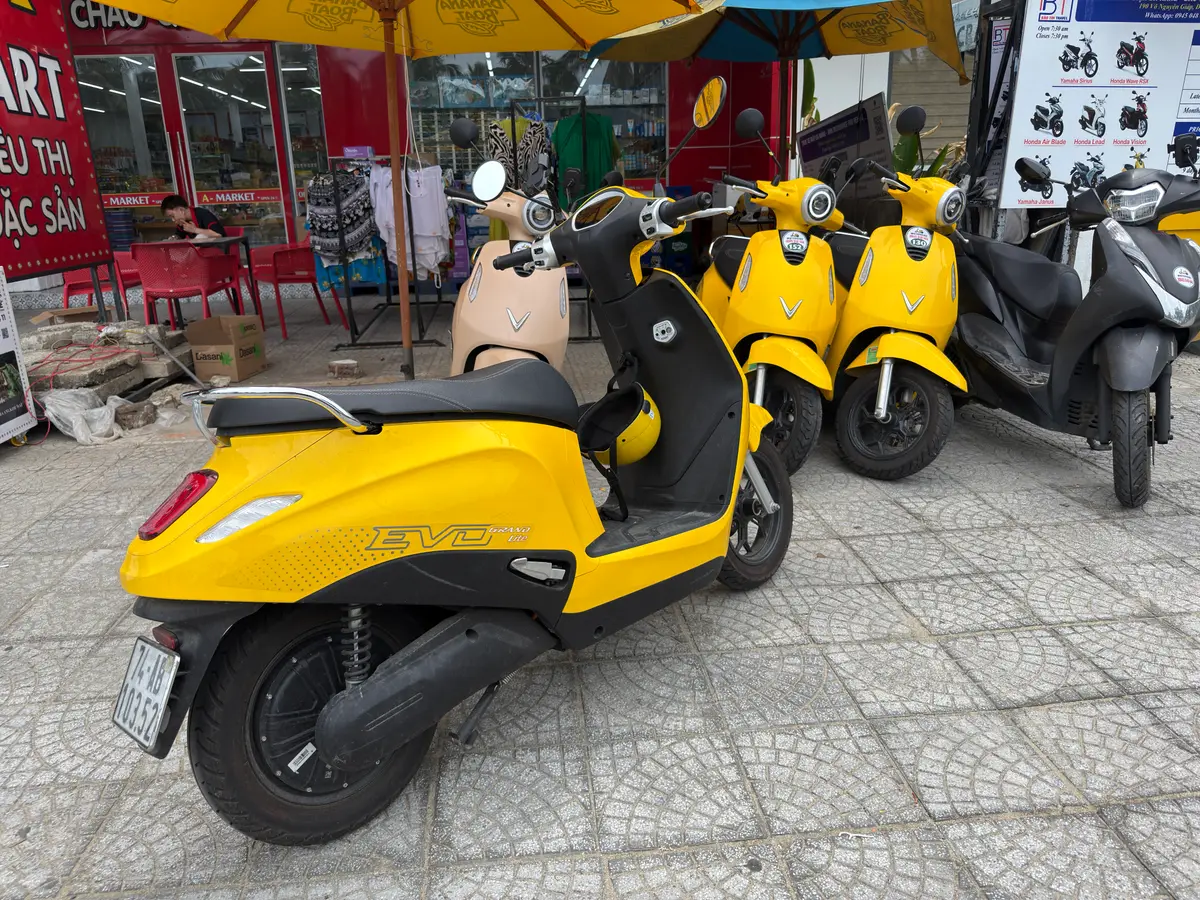 Gelbe Roller und ein schwarzer Roller stehen auf einem gepflasterten Gehweg neben einem Laden mit Sonnenschirmen und einer Tafel mit den angebotenen Modellen.