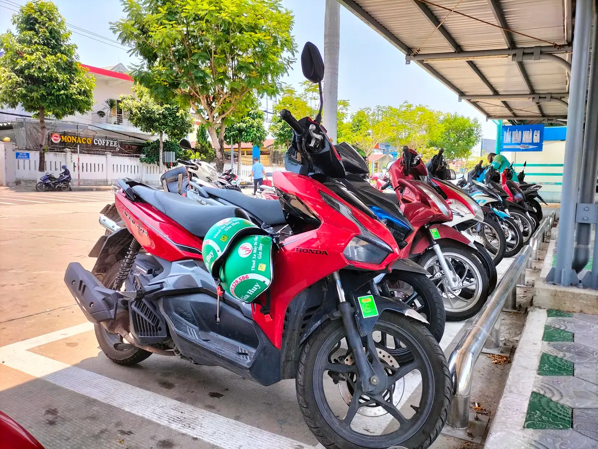 Ein roter Honda-Roller mit einem grünen Helm auf dem Sitz steht auf einem überdachten Stellplatz neben einer Reihe weiterer Fahrzeuge.