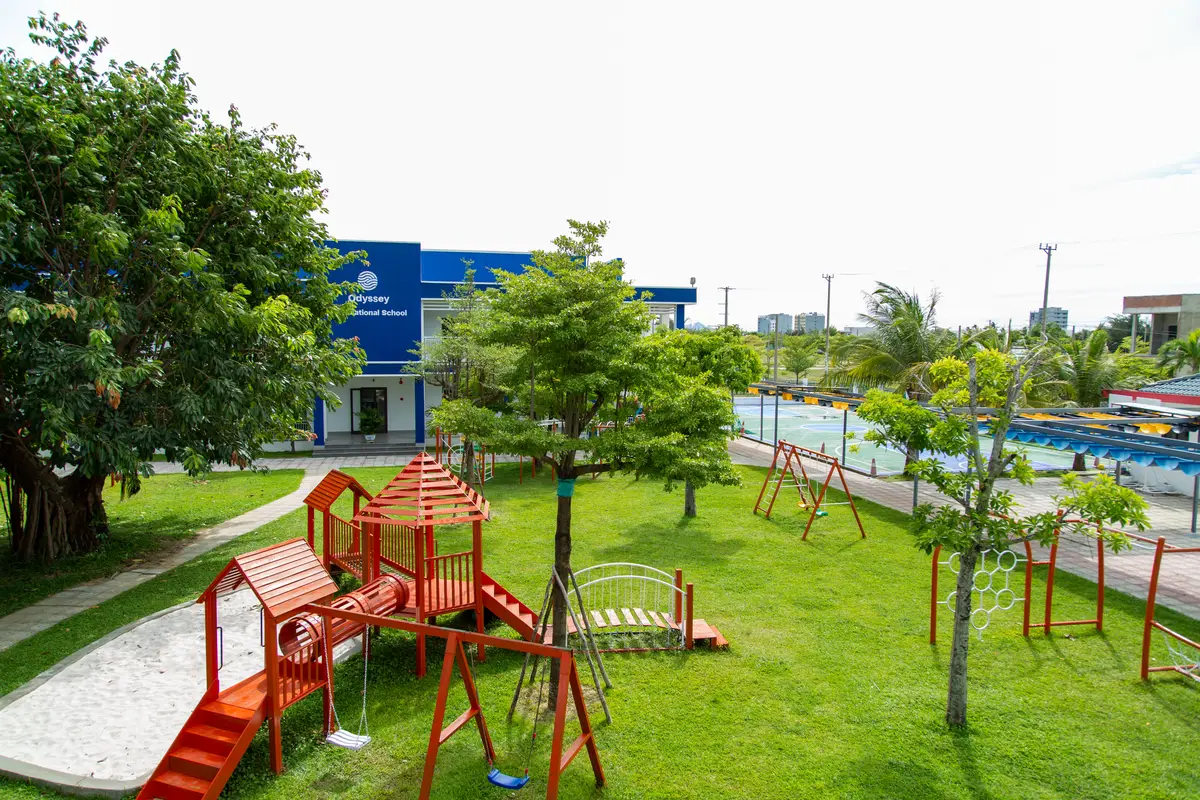 Lawn with a wooden play frame, slides and swings in front of a blue school building with the Odyssey sign