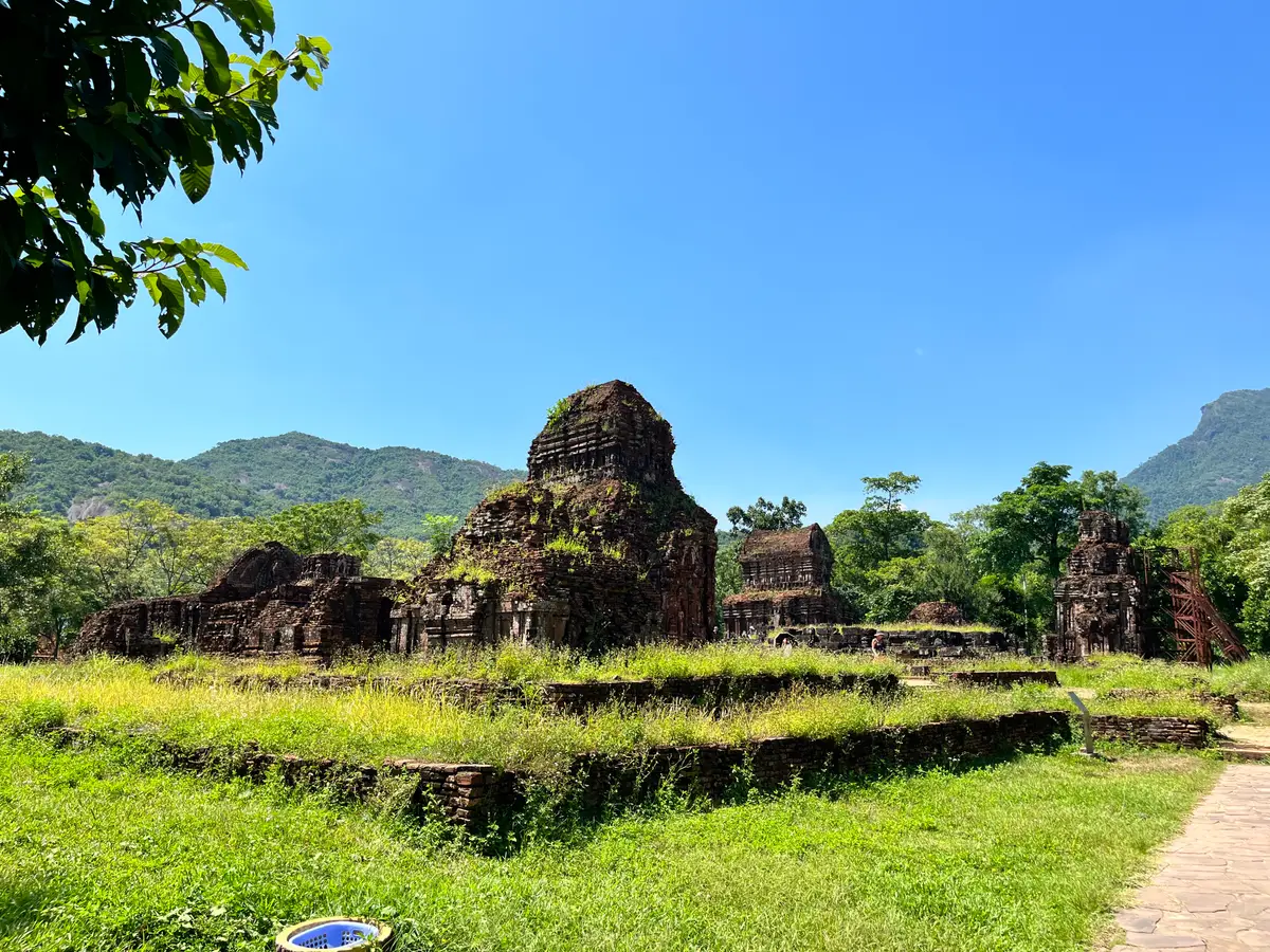 A ruined red-brick tower on a raised platform in a green lawn, with forested mountains behind under a blue sky.