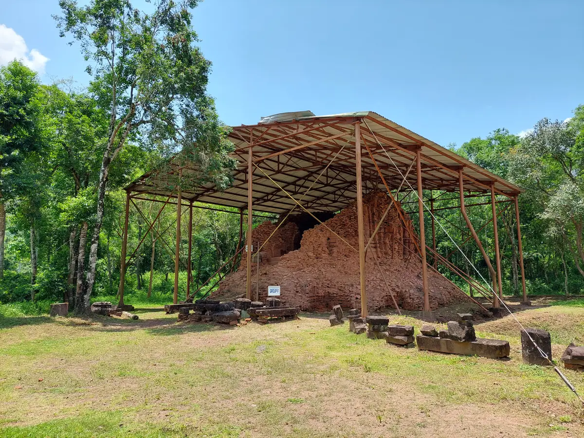 A collapsed brick tower sheltered under a metal roof on posts, with stone fragments laid out on the grass.