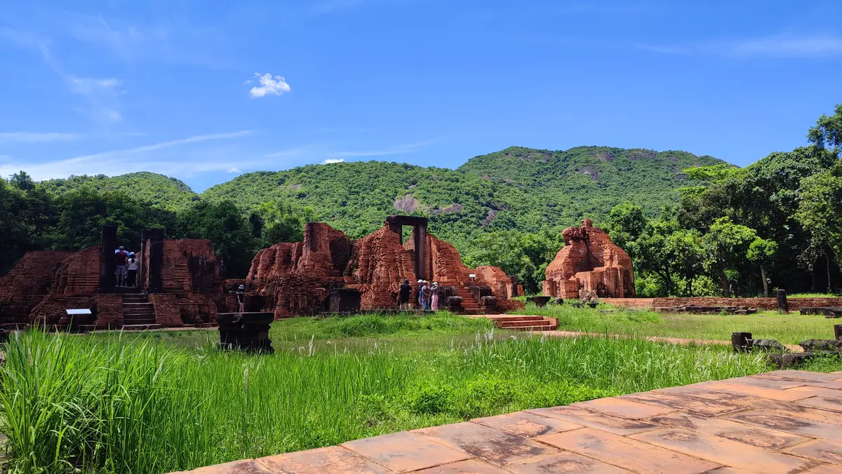 Low red-brick ruins spread across a grassy field, with a paved path in front and green mountains behind.