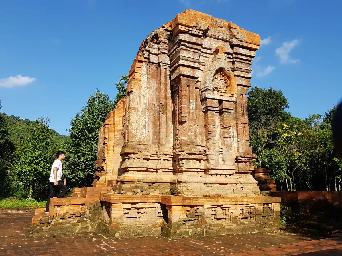 A tall, crumbling brick tower on a stepped base, a visitor standing at its foot for scale.